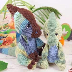 Pu&ograve; includere: Due giocattoli di dinosauro all'uncinetto, uno blu e verde, l'altro verde. Il dinosauro pi&ugrave; grande ha una striscia marrone sulla schiena. Entrambi i dinosauri hanno occhi grandi e sono seduti su una superficie bianca.