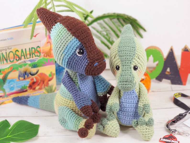 Pu&ograve; includere: Due giocattoli di dinosauro all'uncinetto, uno blu e verde, l'altro verde. Il dinosauro pi&ugrave; grande ha una striscia marrone sulla schiena. Entrambi i dinosauri hanno occhi grandi e sono seduti su una superficie bianca.