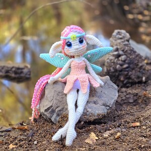 May include: A crocheted fairy doll with pink and white hair, a pink dress, and blue wings sits on a rock.
