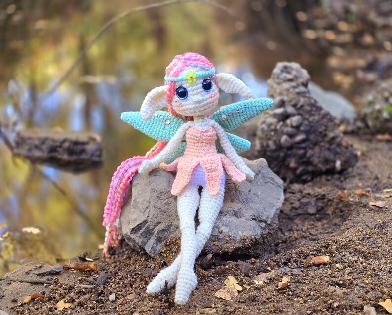 May include: A crocheted fairy doll with pink and white hair, a pink dress, and blue wings sits on a rock.