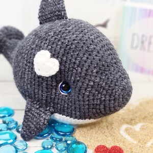 K&ouml;nnte beinhalten: Ein graues geh&auml;keltes Pl&uuml;schwalspielzeug mit einem wei&szlig;en Herzen auf dem Kopf und blauen Augen. Der Wal liegt auf einer Oberfl&auml;che mit blauen Glassteinen und einem Herz, das in den Sand gezeichnet ist.