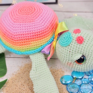 Puede incluir: Un juguete de tortuga marina de crochet con una concha de arco&iacute;ris, un cuerpo verde y ojos grandes. La tortuga lleva un lazo de cinta rosa y arco&iacute;ris.