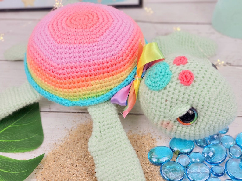 Puede incluir: Un juguete de tortuga marina de crochet con una concha de arco&iacute;ris, un cuerpo verde y ojos grandes. La tortuga lleva un lazo de cinta rosa y arco&iacute;ris.