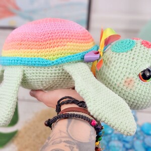 Puede incluir: Un juguete de tortuga de crochet arco&iacute;ris con un caparaz&oacute;n verde y un lazo colorido. La tortuga tiene ojos grandes y negros y una expresi&oacute;n dulce.