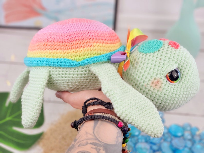 Puede incluir: Un juguete de tortuga de crochet arco&iacute;ris con un caparaz&oacute;n verde y un lazo colorido. La tortuga tiene ojos grandes y negros y una expresi&oacute;n dulce.