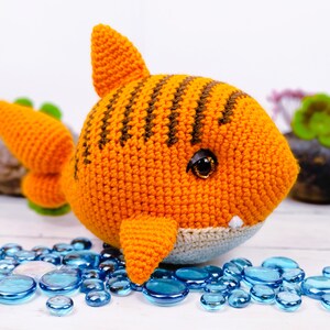 K&ouml;nnte beinhalten: Ein geh&auml;keltes orange-braunes Hai-Spielzeug mit wei&szlig;em Bauch und schwarzen Augen. Der Hai sitzt auf einer wei&szlig;en Oberfl&auml;che mit blauen Glaskieselsteinen.