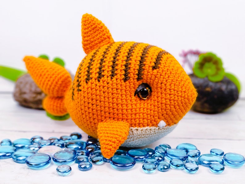 K&ouml;nnte beinhalten: Ein geh&auml;keltes orange-braunes Hai-Spielzeug mit wei&szlig;em Bauch und schwarzen Augen. Der Hai sitzt auf einer wei&szlig;en Oberfl&auml;che mit blauen Glaskieselsteinen.