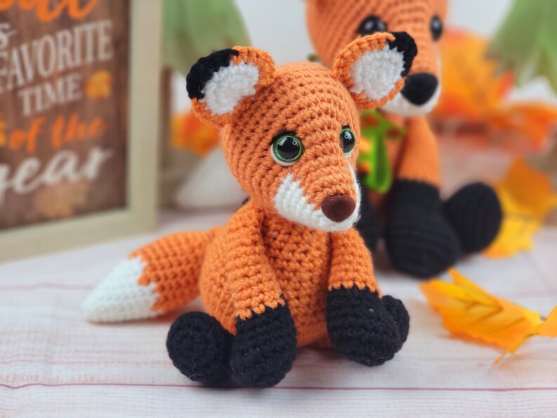 May include: Two crocheted fox plushies, one orange and white with black accents, the other orange with black and white accents. The foxes are sitting on a white surface.