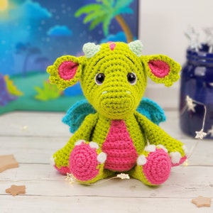 May include: A green crocheted dragon with pink and white accents. The dragon has large eyes and a friendly expression. It is sitting on a white surface.