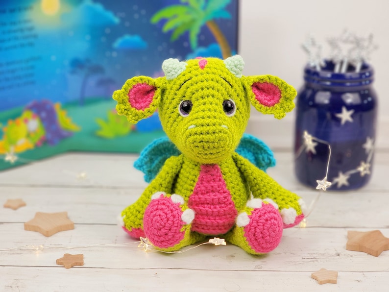 May include: A green crocheted dragon with pink and white accents. The dragon has large eyes and a friendly expression. It is sitting on a white surface.
