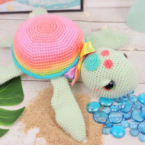 Puede incluir: Un juguete de tortuga marina de crochet con una concha de arco&iacute;ris y un cuerpo verde. La tortuga tiene ojos grandes y negros y detalles rosas en su caparaz&oacute;n. El juguete est&aacute; sentado en una superficie que parece arena.