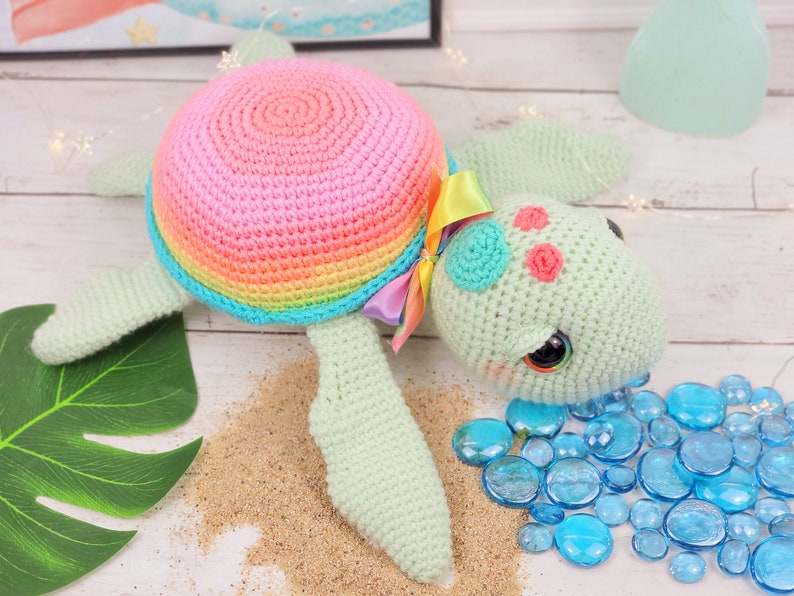 Puede incluir: Un juguete de tortuga marina de crochet con una concha de arco&iacute;ris y un cuerpo verde. La tortuga tiene ojos grandes y negros y detalles rosas en su caparaz&oacute;n. El juguete est&aacute; sentado en una superficie que parece arena.