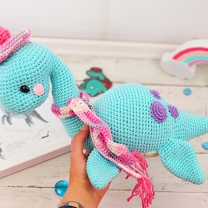 May include: A crocheted blue dinosaur toy wearing a pink and white striped hat and scarf. The dinosaur has purple spots on its back.