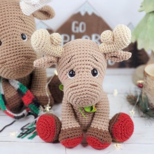 May include: Two crocheted moose toys, one brown with a red and green scarf and the other brown with red feet. The moose are sitting on a white surface with a wooden sign that says "Go Explore" behind them.