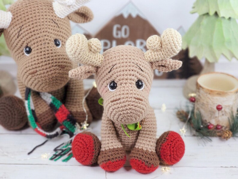 May include: Two crocheted moose toys, one brown with a red and green scarf and the other brown with red feet. The moose are sitting on a white surface with a wooden sign that says "Go Explore" behind them.