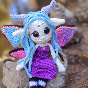 May include: A crocheted fairy doll with blue hair, purple dress, and iridescent wings. The doll is sitting on a rock.