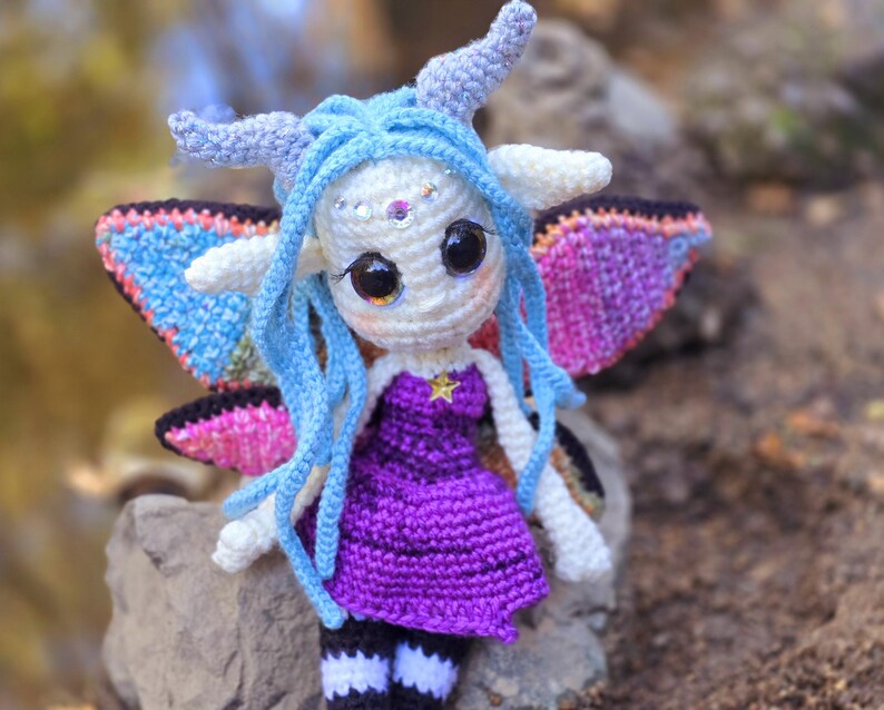 May include: A crocheted fairy doll with blue hair, purple dress, and iridescent wings. The doll is sitting on a rock.