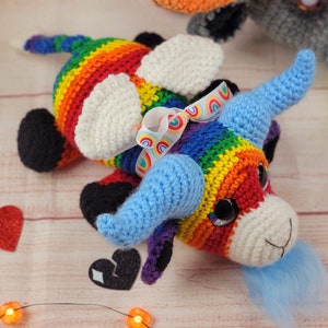 Peut inclure: Unicorn crochet fait &agrave; la main aux couleurs de l'arc-en-ciel. La licorne a des cornes bleues, des taches blanches et une crini&egrave;re bleue duveteuse. Un n&oelig;ud en ruban arc-en-ciel orne la t&ecirc;te. Le jouet est pos&eacute; sur une surface en bois clair.