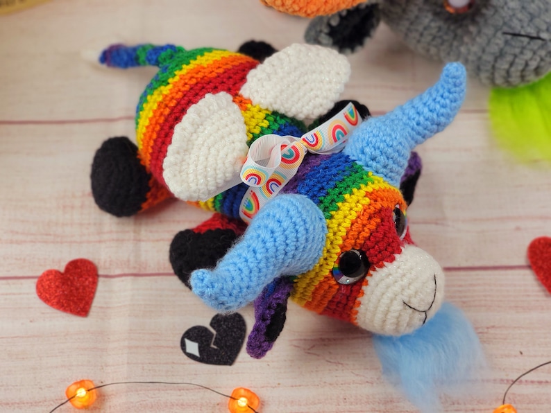 Peut inclure: Unicorn crochet fait &agrave; la main aux couleurs de l'arc-en-ciel. La licorne a des cornes bleues, des taches blanches et une crini&egrave;re bleue duveteuse. Un n&oelig;ud en ruban arc-en-ciel orne la t&ecirc;te. Le jouet est pos&eacute; sur une surface en bois clair.