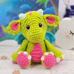 Puede incluir: Un drag&oacute;n de crochet verde con orejas, alas y pies rosas. El drag&oacute;n tiene cuernos blancos y ojos negros.