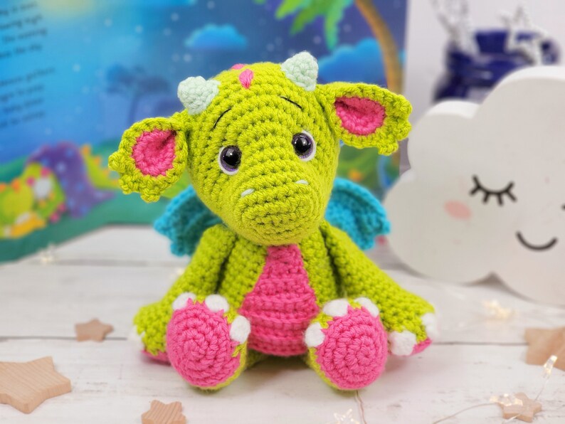 Puede incluir: Un drag&oacute;n de crochet verde con orejas, alas y pies rosas. El drag&oacute;n tiene cuernos blancos y ojos negros.