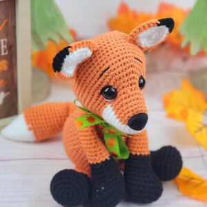 May include: A crocheted orange fox with black eyes, a white nose, and black feet. The fox is wearing a green ribbon with a leaf design. The fox is sitting on a white surface with fall leaves in the background.