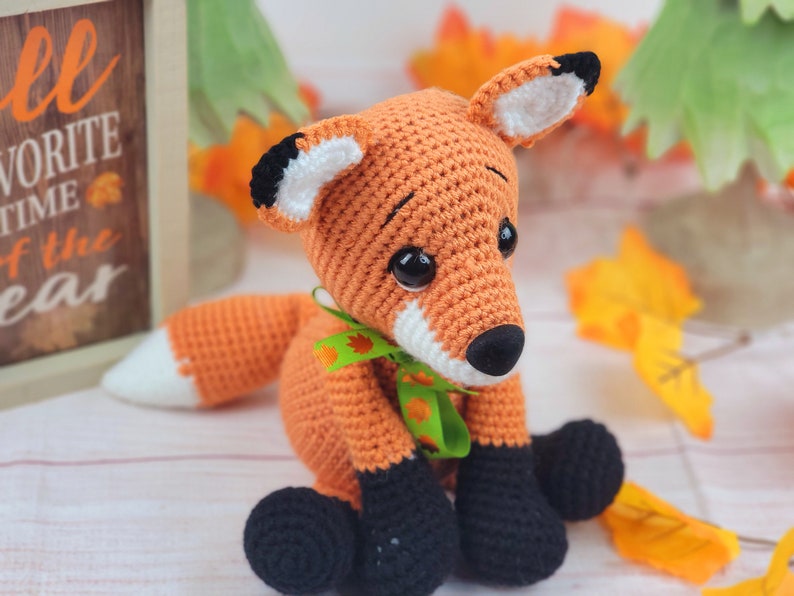 May include: A crocheted orange fox with black eyes, a white nose, and black feet. The fox is wearing a green ribbon with a leaf design. The fox is sitting on a white surface with fall leaves in the background.