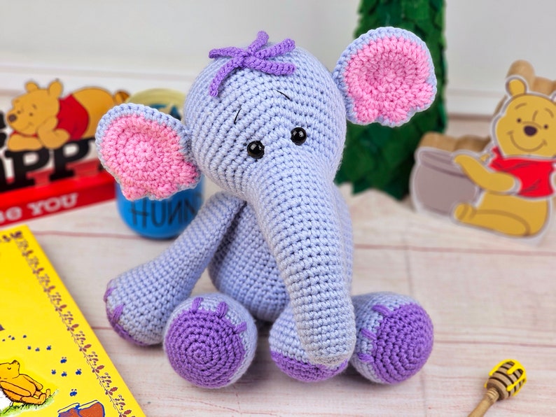 May include: A crocheted purple and pink elephant toy with a purple flower on its head. The elephant is sitting on a wooden surface.