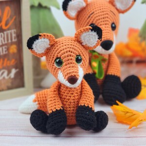 May include: Two crocheted fox plushies, one orange with black accents and the other orange with green accents. The foxes are sitting on a light wood surface with fall leaves scattered around them. A sign behind the foxes reads "Fall is my favorite time of the year."