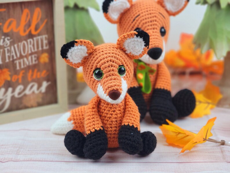 May include: Two crocheted fox plushies, one orange with black accents and the other orange with green accents. The foxes are sitting on a light wood surface with fall leaves scattered around them. A sign behind the foxes reads "Fall is my favorite time of the year."