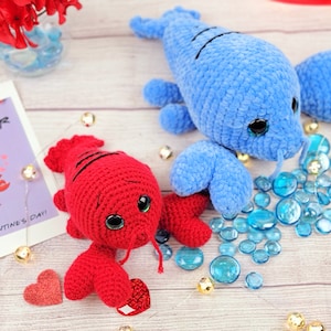 Puede incluir: Dos peluches de langosta de ganchillo, uno rojo y otro azul, con grandes ojos saltones. También hay una tarjeta de San Valentín con una ilustración de langosta y el texto "You're my Lobster", junto con cuentas de vidrio decorativas.