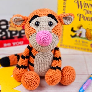 May include: A crocheted stuffed toy tiger with orange and black stripes, a pink nose, and black eyes. The toy is sitting on a white surface with a yellow sunflower behind it.