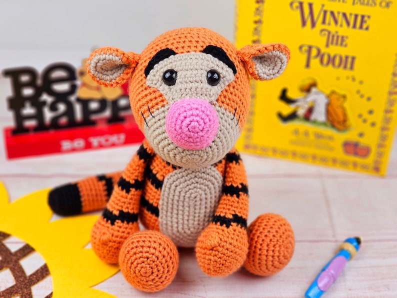 May include: A crocheted stuffed toy tiger with orange and black stripes, a pink nose, and black eyes. The toy is sitting on a white surface with a yellow sunflower behind it.