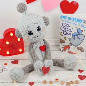 May include: A handmade gray crochet robot doll with a red heart on its chest and holding a red heart. A book titled "Ready-to-Read The adventures of Otto, Go, Otto, Go!" is visible. Decorative hearts and the words "Be Mine" are also present.