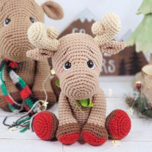 May include: Two crocheted moose toys, one wearing a red and green scarf, sitting on a white wooden surface. The moose on the right has red booties.