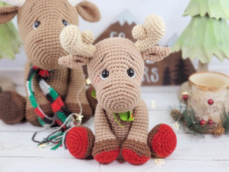May include: Two crocheted moose toys, one wearing a red and green scarf, sitting on a white wooden surface. The moose on the right has red booties.