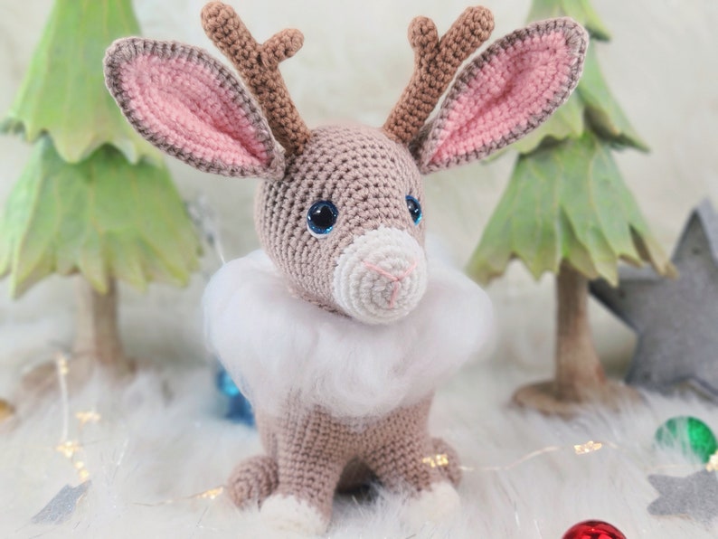 May include: A handmade crochet reindeer with a light brown body, pink-lined ears, and brown antlers. It has blue eyes and a white fluffy collar. The reindeer is a festive decoration, perfect for the holiday season.