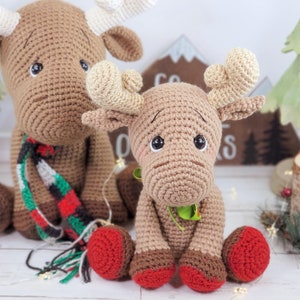 May include: Two crocheted brown moose with red and green accents. The larger moose is wearing a red and green plaid scarf. The smaller moose is sitting in front of the larger moose.