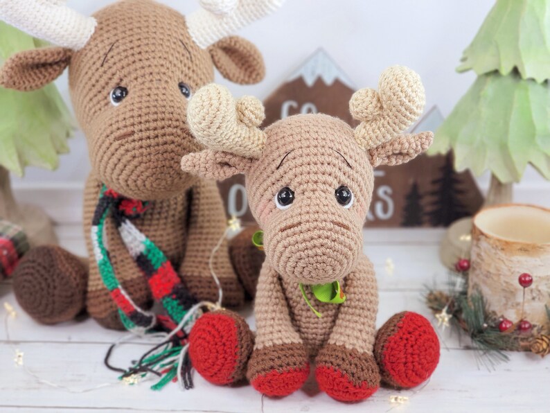 May include: Two crocheted brown moose with red and green accents. The larger moose is wearing a red and green plaid scarf. The smaller moose is sitting in front of the larger moose.