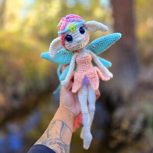 May include: A crocheted fairy doll with pink, blue, and white yarn. The fairy has large blue eyes, a pink dress, and blue wings with iridescent glitter.