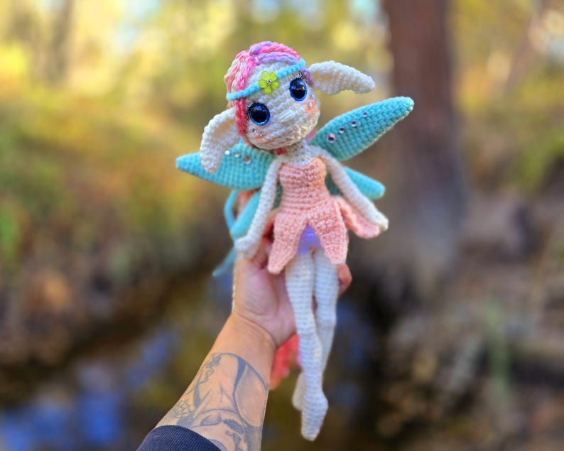 May include: A crocheted fairy doll with pink, blue, and white yarn. The fairy has large blue eyes, a pink dress, and blue wings with iridescent glitter.