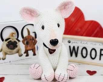 May include: White crocheted bull terrier dog with pink ears and paws. The dog is wearing a black collar with a red heart-shaped tag that says "Champ". The dog is sitting on a white wooden surface in front of a red heart and a sign that says "You had me at woof."