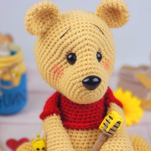 K&ouml;nnte beinhalten: Ein geh&auml;keltes Winnie Puuh-B&auml;ren-Spielzeug, das ein rotes Hemd tr&auml;gt und einen Honigl&ouml;ffel h&auml;lt. Der B&auml;r hat eine braune Nase und schwarze Augen.