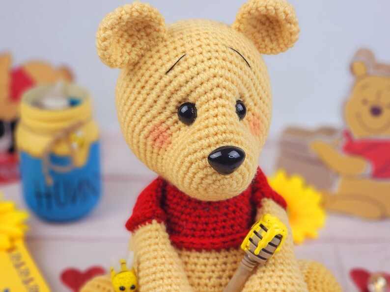 K&ouml;nnte beinhalten: Ein geh&auml;keltes Winnie Puuh-B&auml;ren-Spielzeug, das ein rotes Hemd tr&auml;gt und einen Honigl&ouml;ffel h&auml;lt. Der B&auml;r hat eine braune Nase und schwarze Augen.