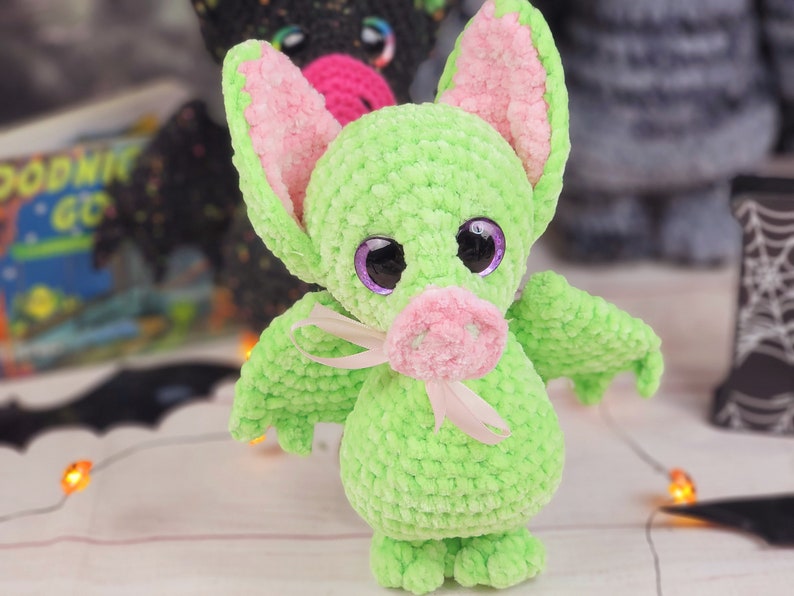 May include: A green crocheted bat with pink ears and a pink nose. The bat has large purple eyes and a white ribbon around its neck.