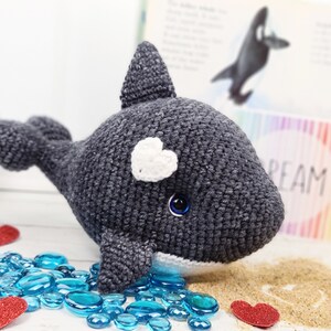 K&ouml;nnte beinhalten: Ein geh&auml;keltes graues und wei&szlig;es Orca-Wal-Pl&uuml;schtier mit einem wei&szlig;en Herzen auf dem Kopf und blauen Augen. Das Spielzeug sitzt auf einer wei&szlig;en Oberfl&auml;che mit blauen Glassteinen und roten Herzen.