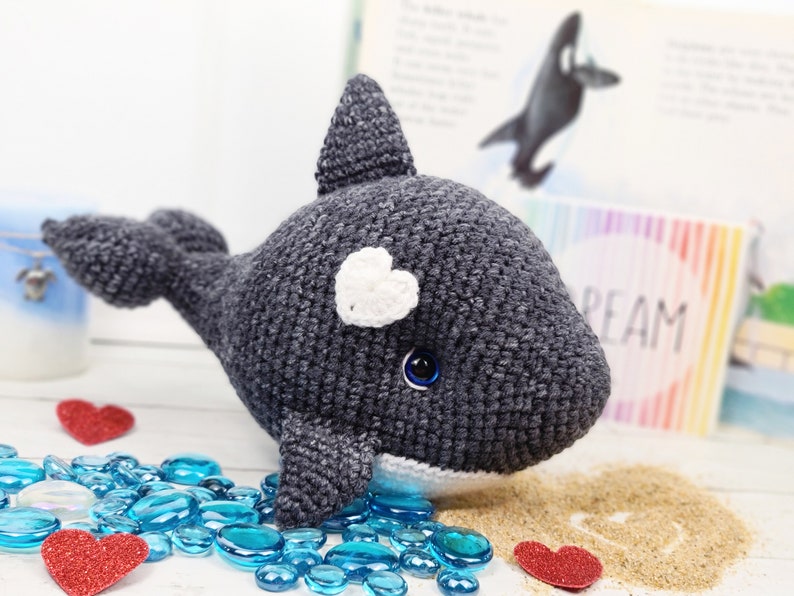 K&ouml;nnte beinhalten: Ein geh&auml;keltes graues und wei&szlig;es Orca-Wal-Pl&uuml;schtier mit einem wei&szlig;en Herzen auf dem Kopf und blauen Augen. Das Spielzeug sitzt auf einer wei&szlig;en Oberfl&auml;che mit blauen Glassteinen und roten Herzen.