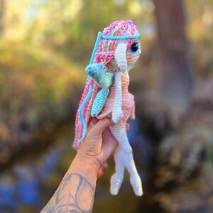 May include: A crocheted fairy doll with pink, blue, and white yarn. The doll has a long, flowing skirt and wings. The doll is holding a small, blue flower.