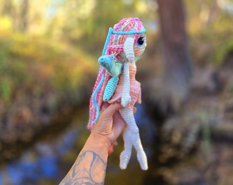 May include: A crocheted fairy doll with pink, blue, and white yarn. The doll has a long, flowing skirt and wings. The doll is holding a small, blue flower.