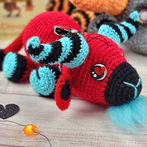 Peut inclure: Une cr&eacute;ature rouge faite &agrave; la main au crochet avec des accents noirs et bleus. Le jouet en peluche a un museau noir, une touffe bleue et un &oelig;il bouton avec un c&oelig;ur bris&eacute;. D'autres jouets au crochet sont en arri&egrave;re-plan.